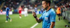 Un arbitre masculin de football, sifflet à la main, observant une action sur le bord du terrain dans une ambiance de match dynamique, avec pelouse verdoyante et stade en arrière-plan.