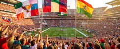 Vue grand angle d’un stade de football nord-américain rempli de supporters, avec trois grands drapeaux des pays hôtes flottant au-dessus des tribunes pendant un match international estival.