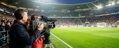 Un homme et une femme cadreurs filment un match de football depuis la ligne de touche dans un stade éclairé, avec des caméras professionnelles et des tribunes animées.